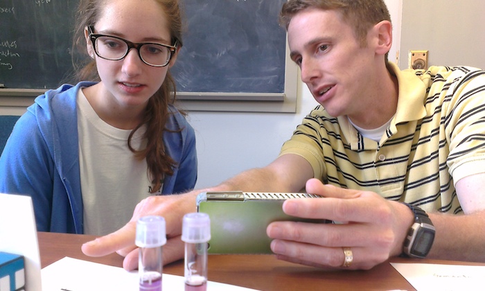 A student researcher and instructor compare notes in front of a pair of test tubes as one of them holds a cell phone