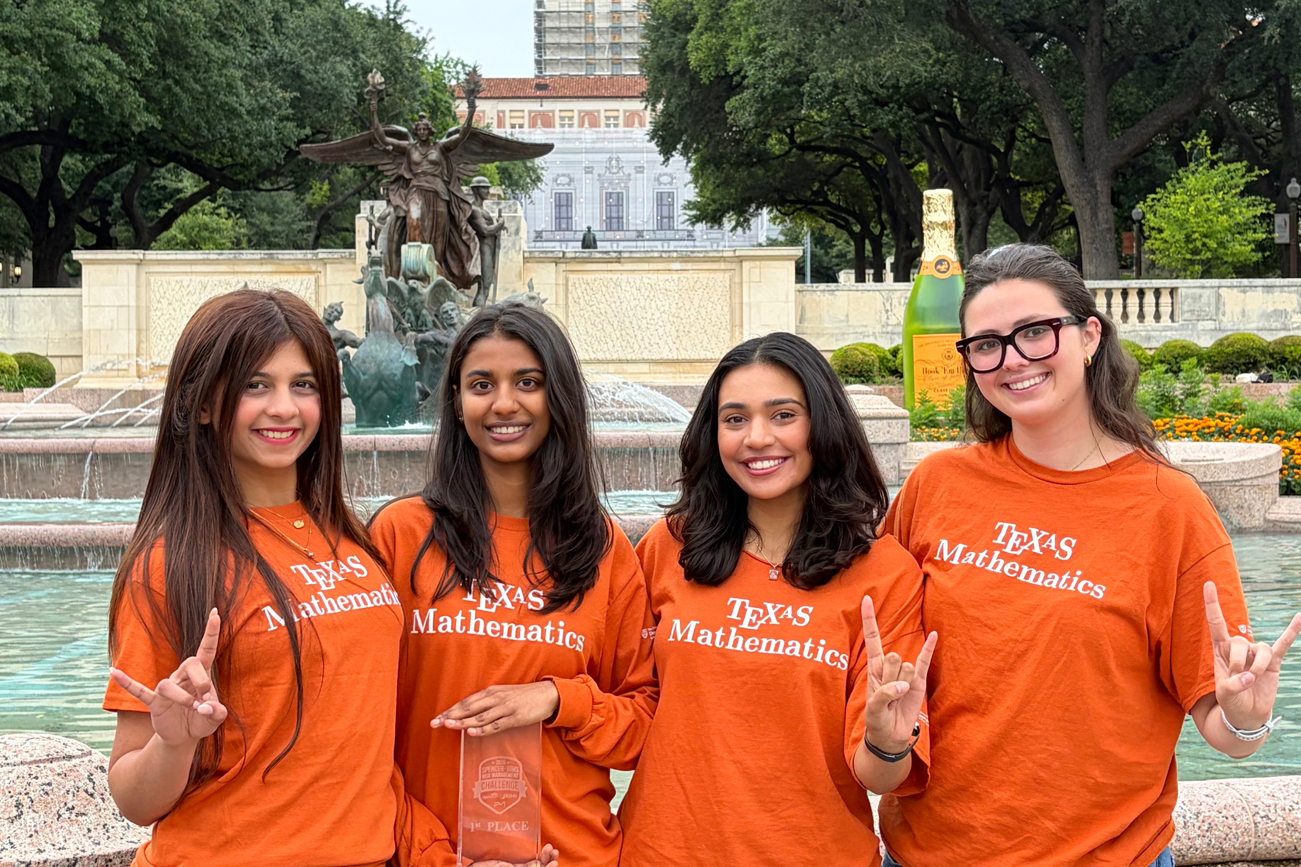 Four young women in Texas Mathematics burnt orange shirts pose before the UT Tower with the hook 'em horns sign.