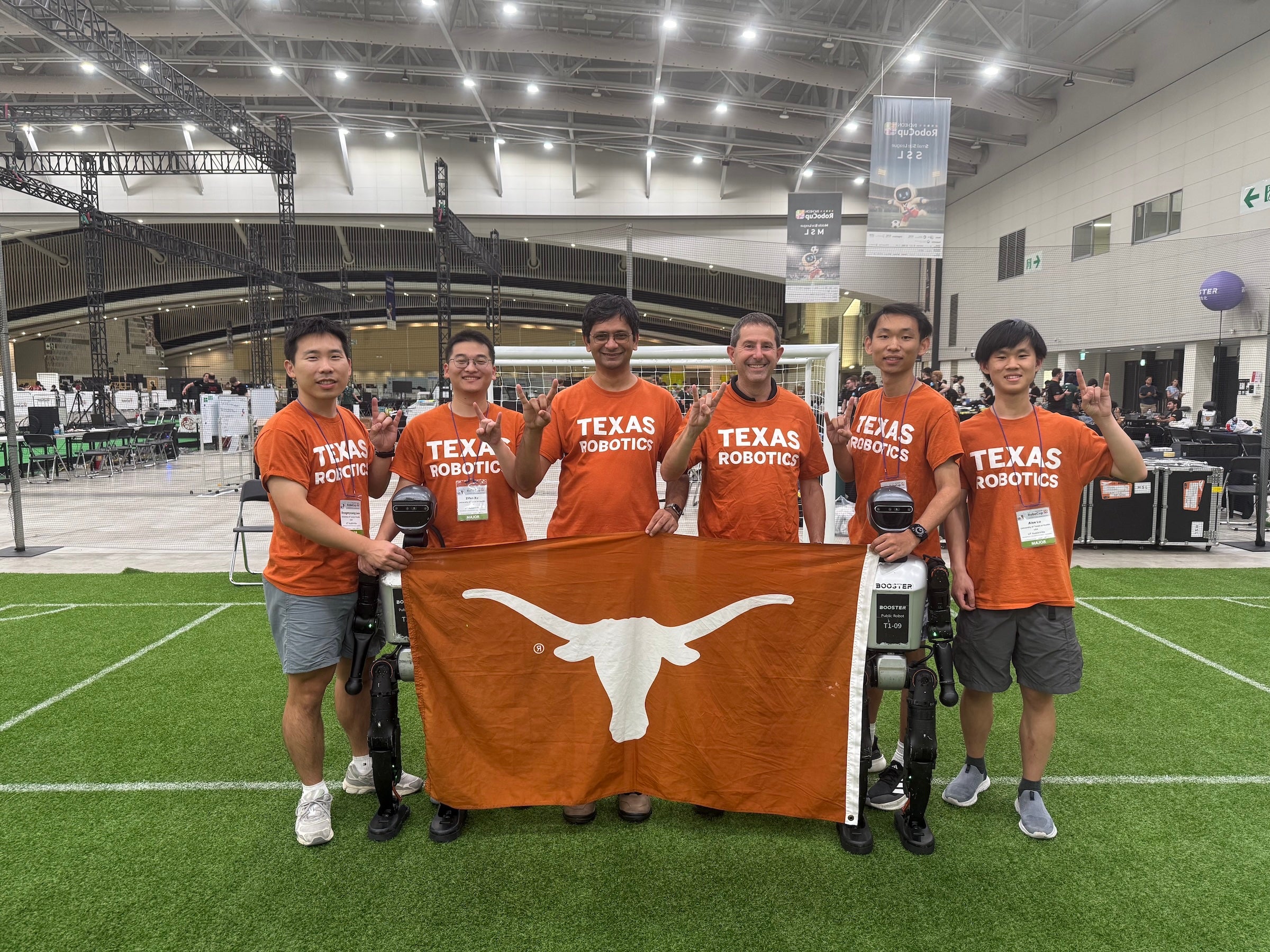 Six men in Texas Robotics t-shirts stand behind a flag with the Longhorn silhouette and a pair of humanoid robots in a convention hall on an artificial soccer field.