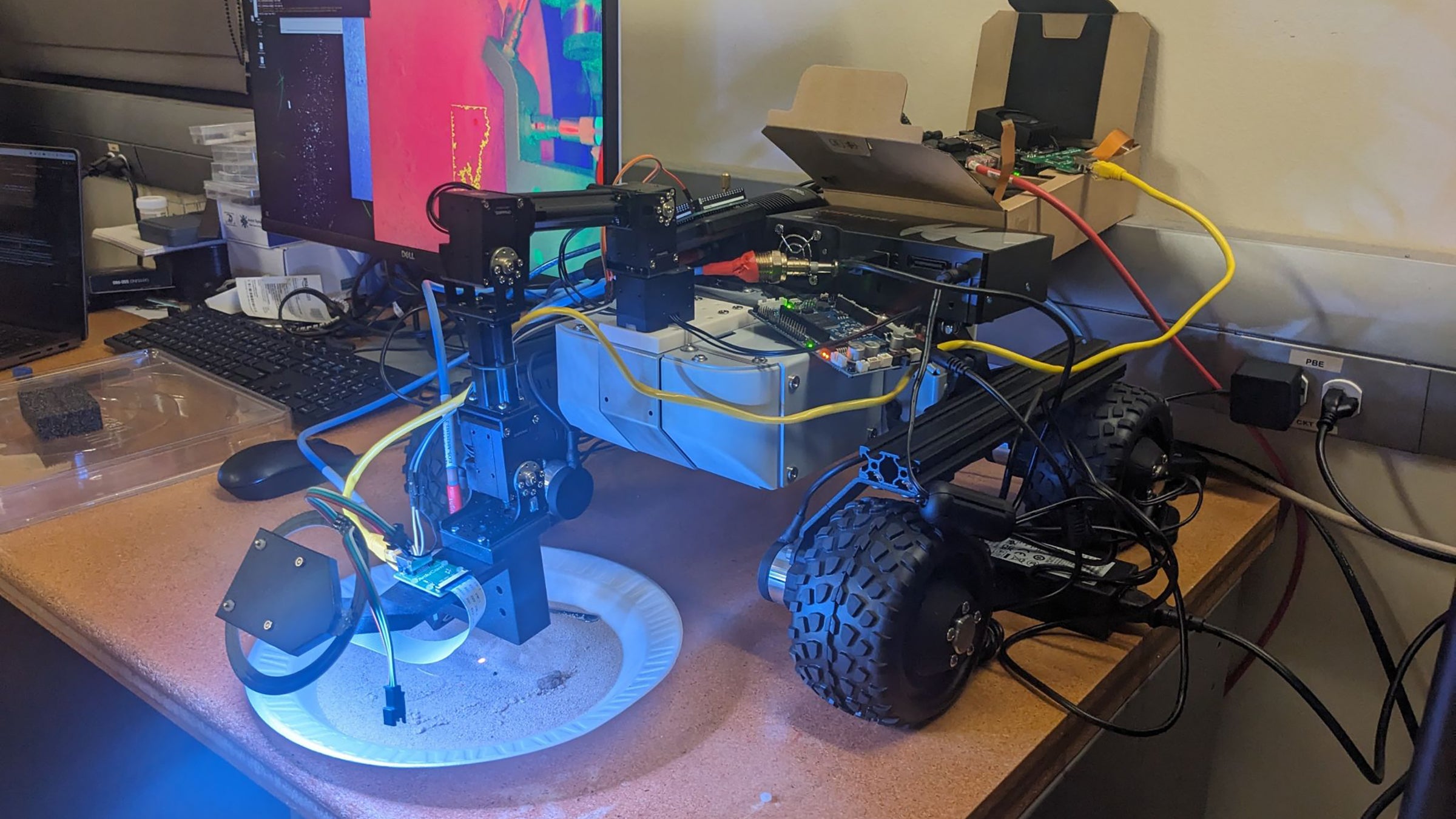 A toaster-sized robot sits on a table with and mechanical arm hovering over a dish of sand. A bluish light points onto a spot on the sand for analysis.