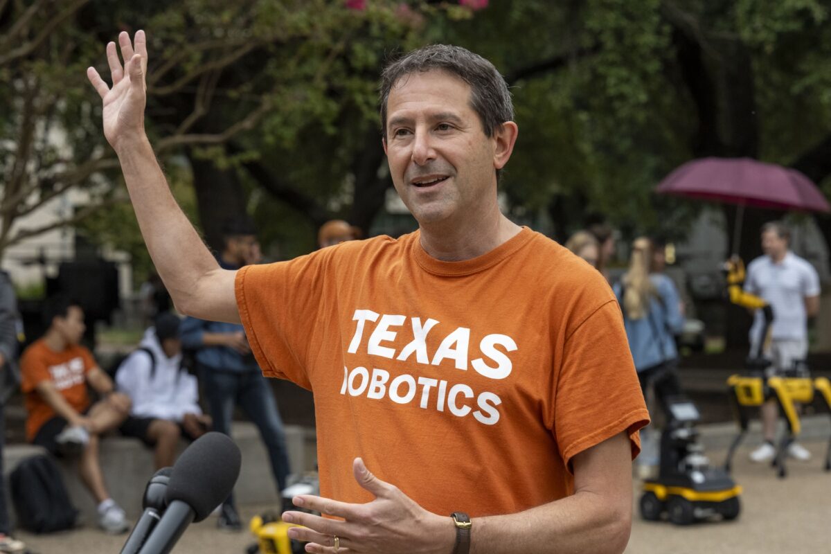 A man in a Texas Robotics t-shirt speaks at a mic, gesturing with his hands as robots and people interact in the background.