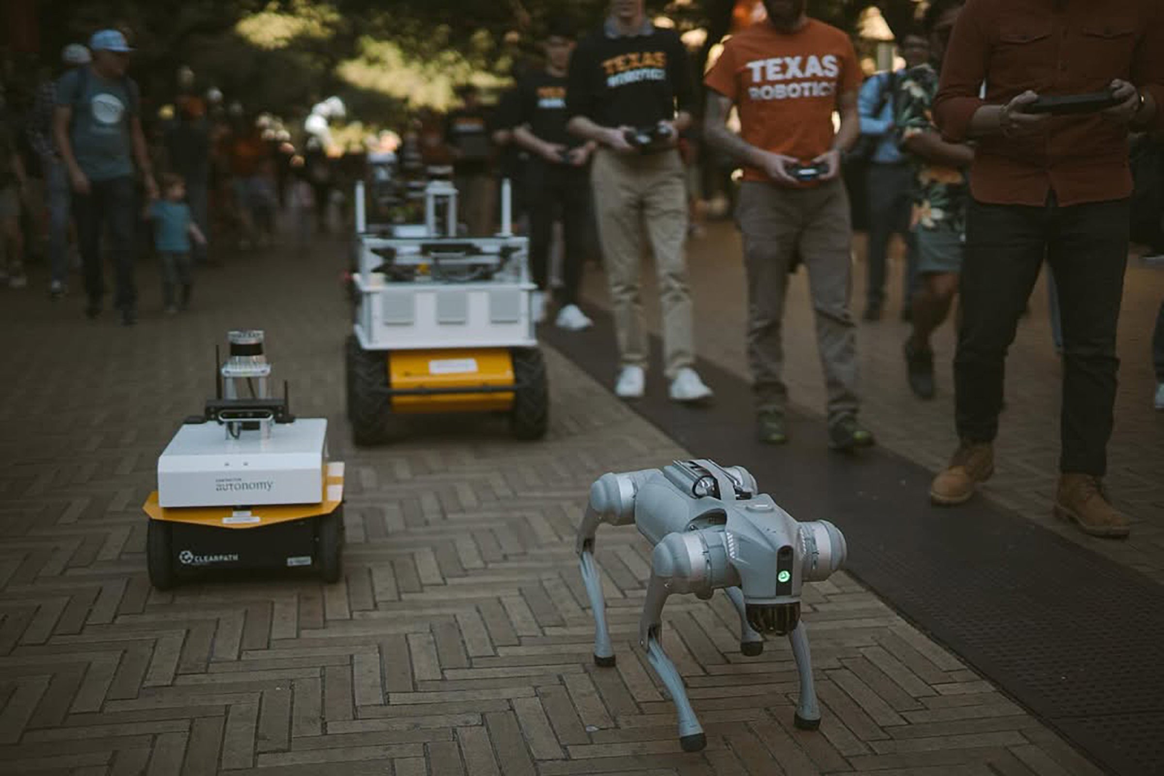 Three robots of varying design move along a brick walkway next to their human controllers.