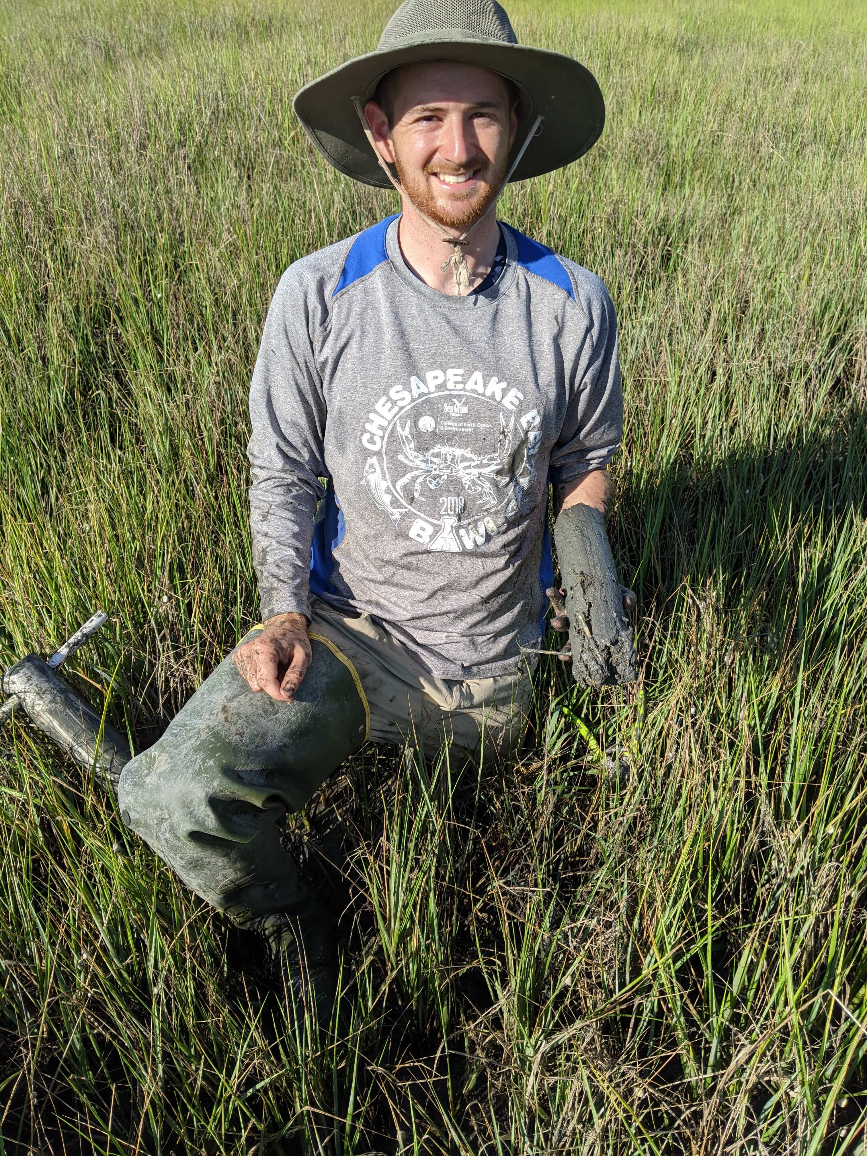 A bearded man in a sun hat smiles in a marshy area while mud cakes his clothes and equipment.