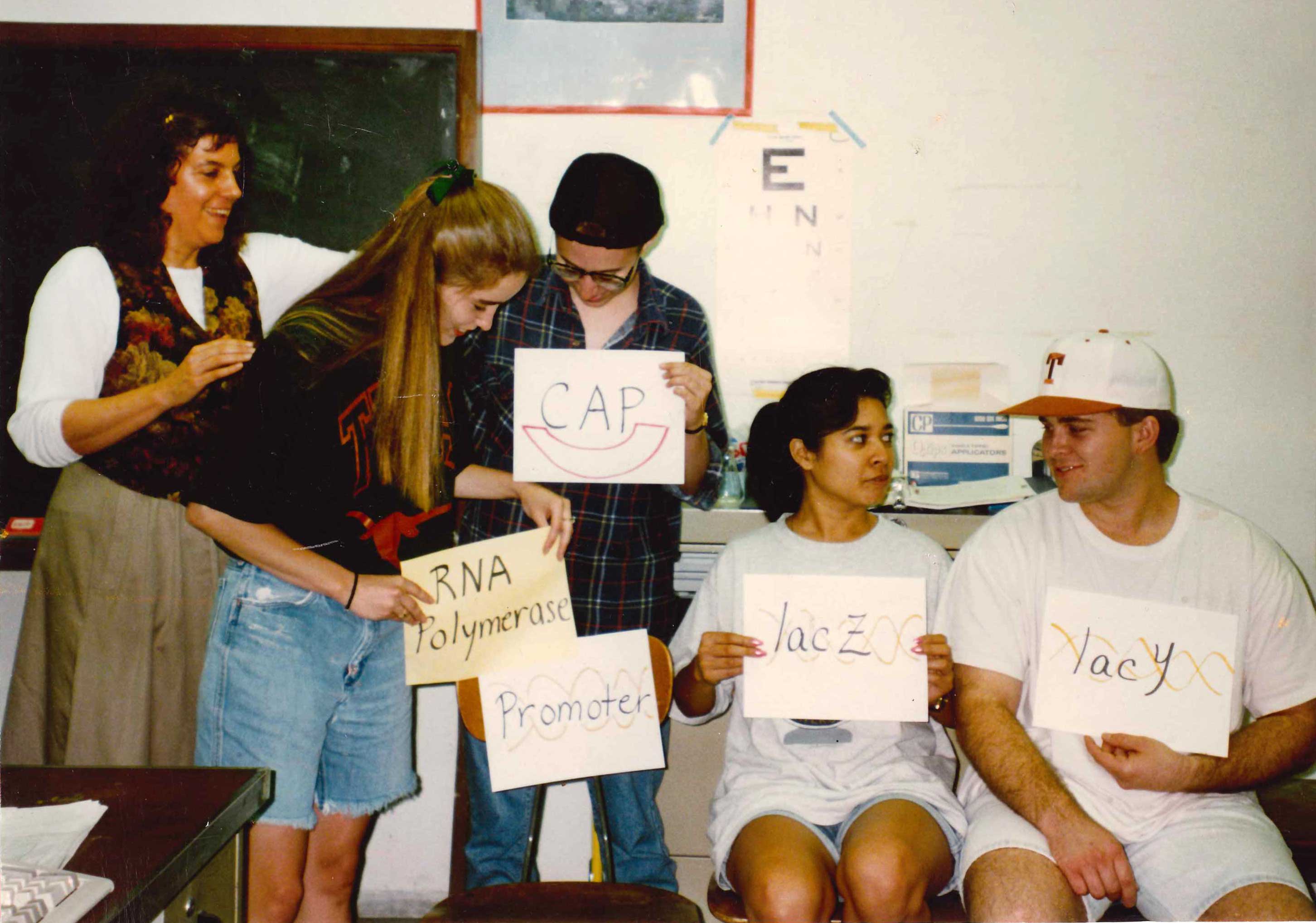Students at the front of a classroom perform a skit holding signs that identify biological molecules, such as lacY, lacZ, RNA polymerase, promoter and CAP. A professor stands next to them.