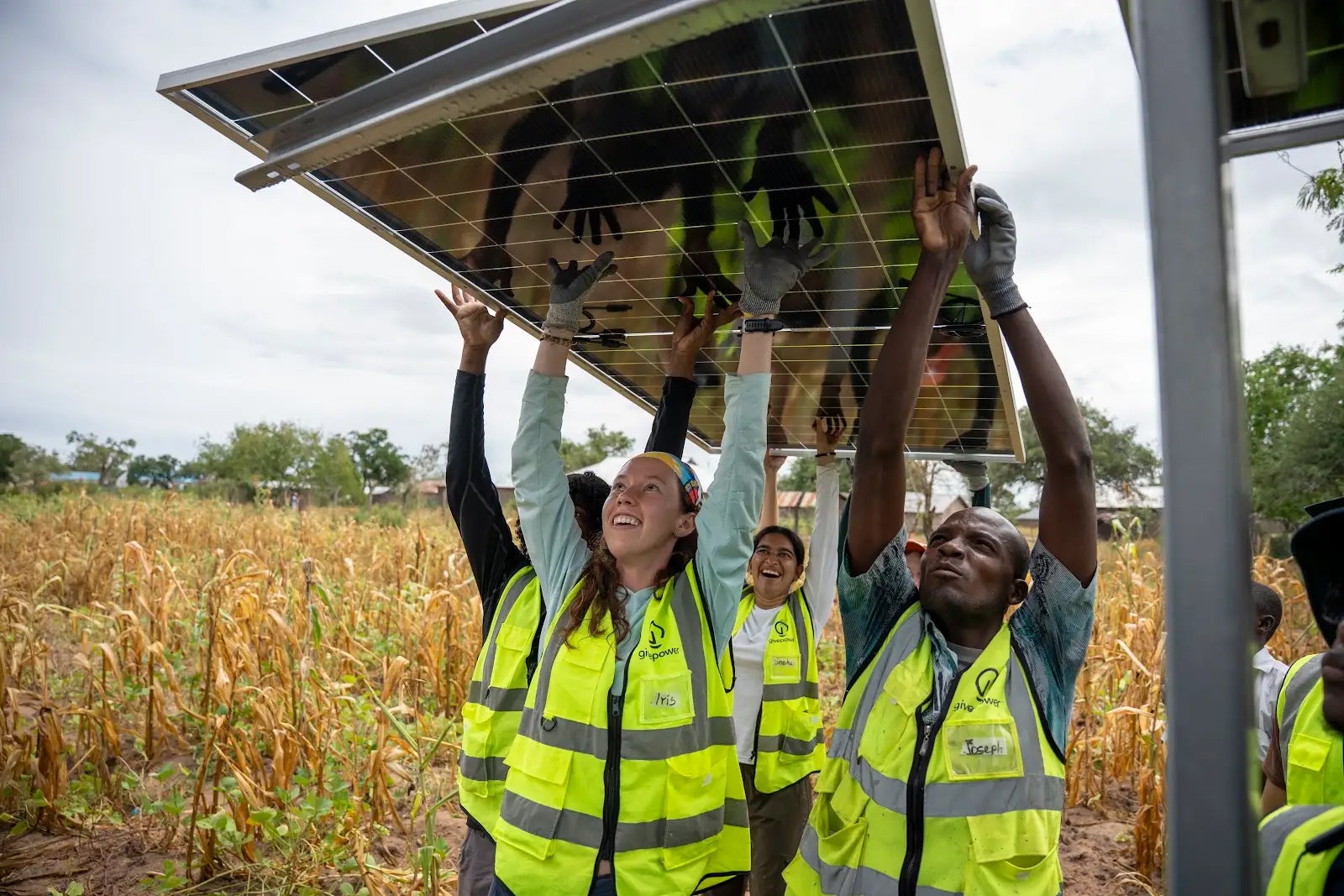 Several people wearing yellow safety vests carry a solar panel above their heads