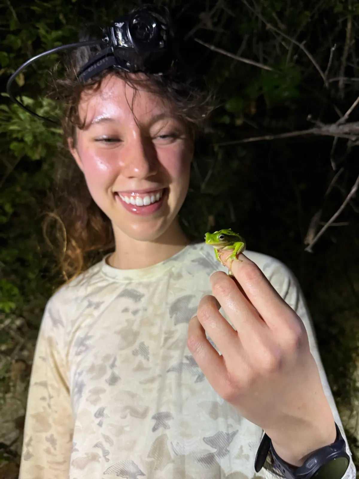 A woman standing in a wooded area and wearing a headlamp holds a frog in her hand
