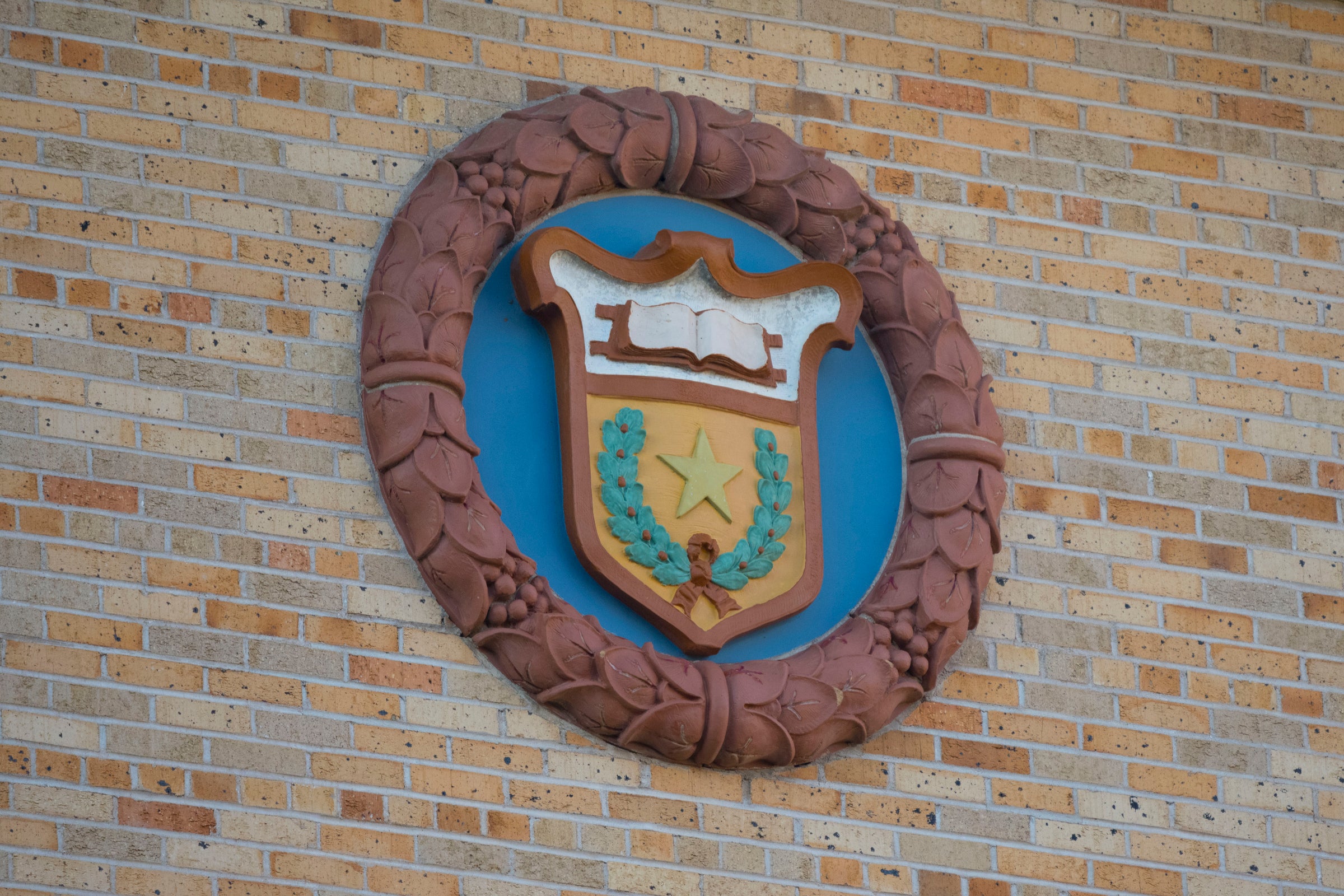 Seal of the University of Texas at Austin, a crest with a book, garland, star within a circle, against the bricks of Painter Hall