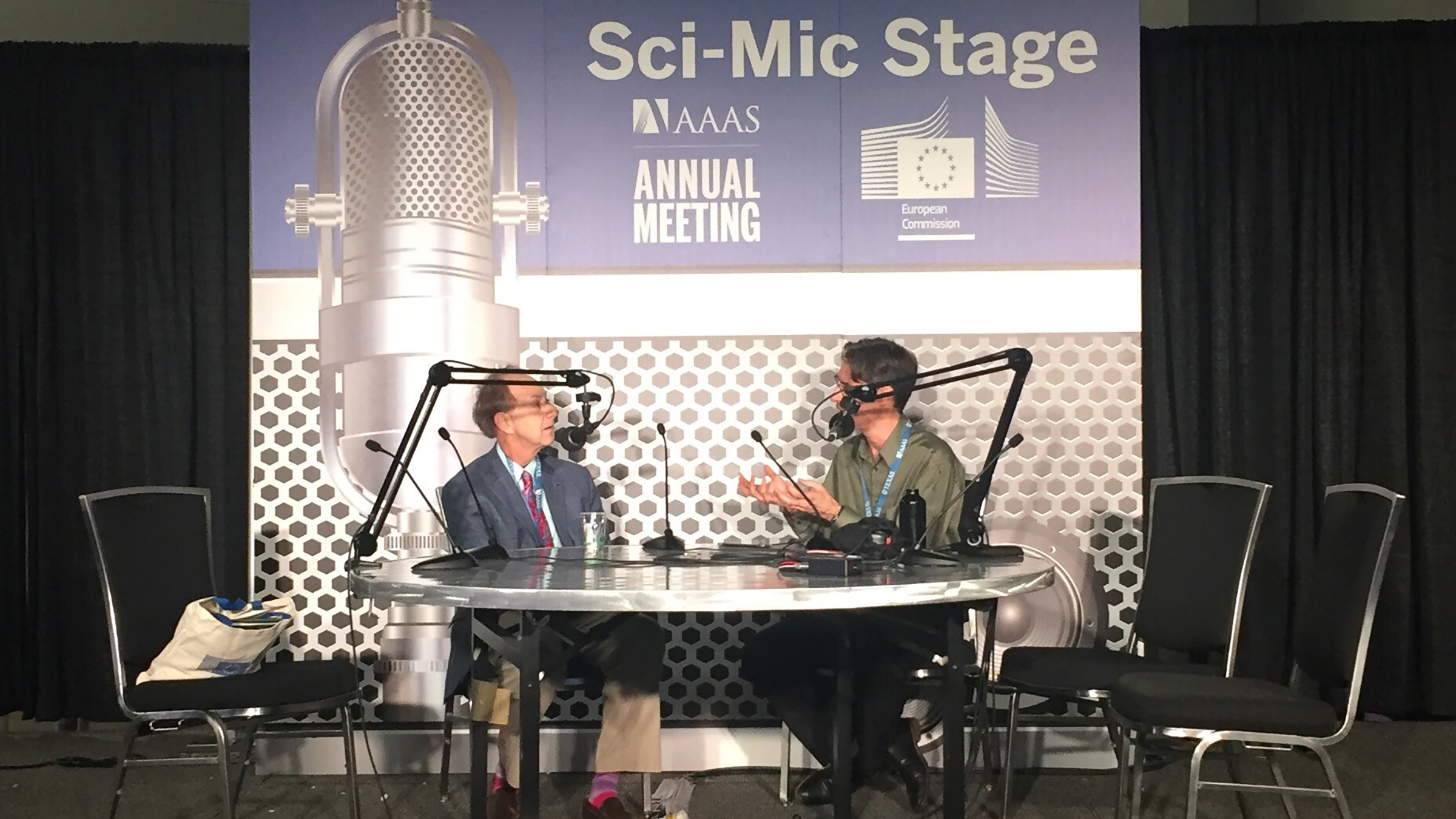 Two men sit at a desk with microphones, a banner behind them reads "Sci-Mic Stage"