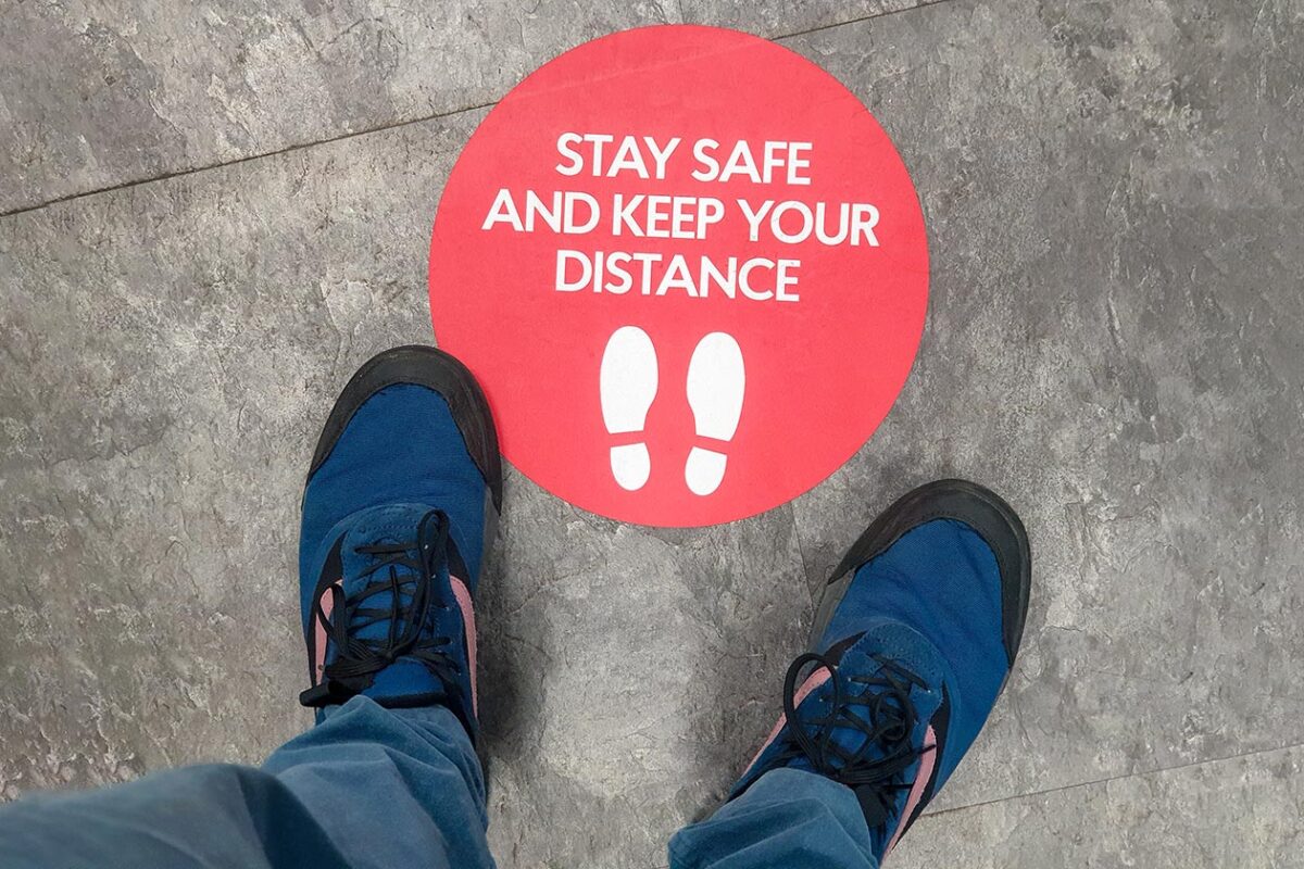 Image shows a person's feet in tennis shoes standing near a red circle sticker that reads stay safe and keep your distance