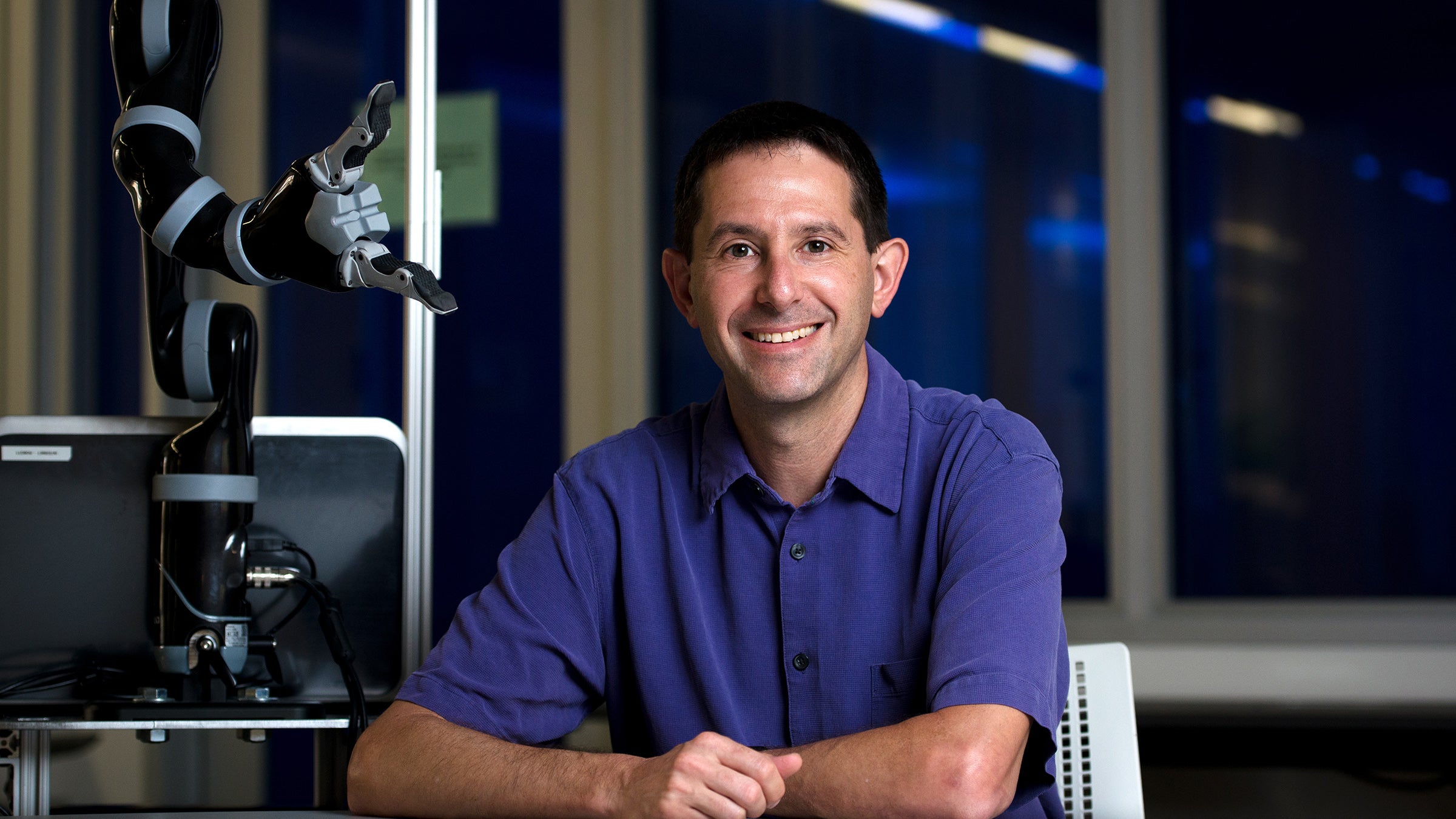 A man leans against a desk beside a robotic arm 