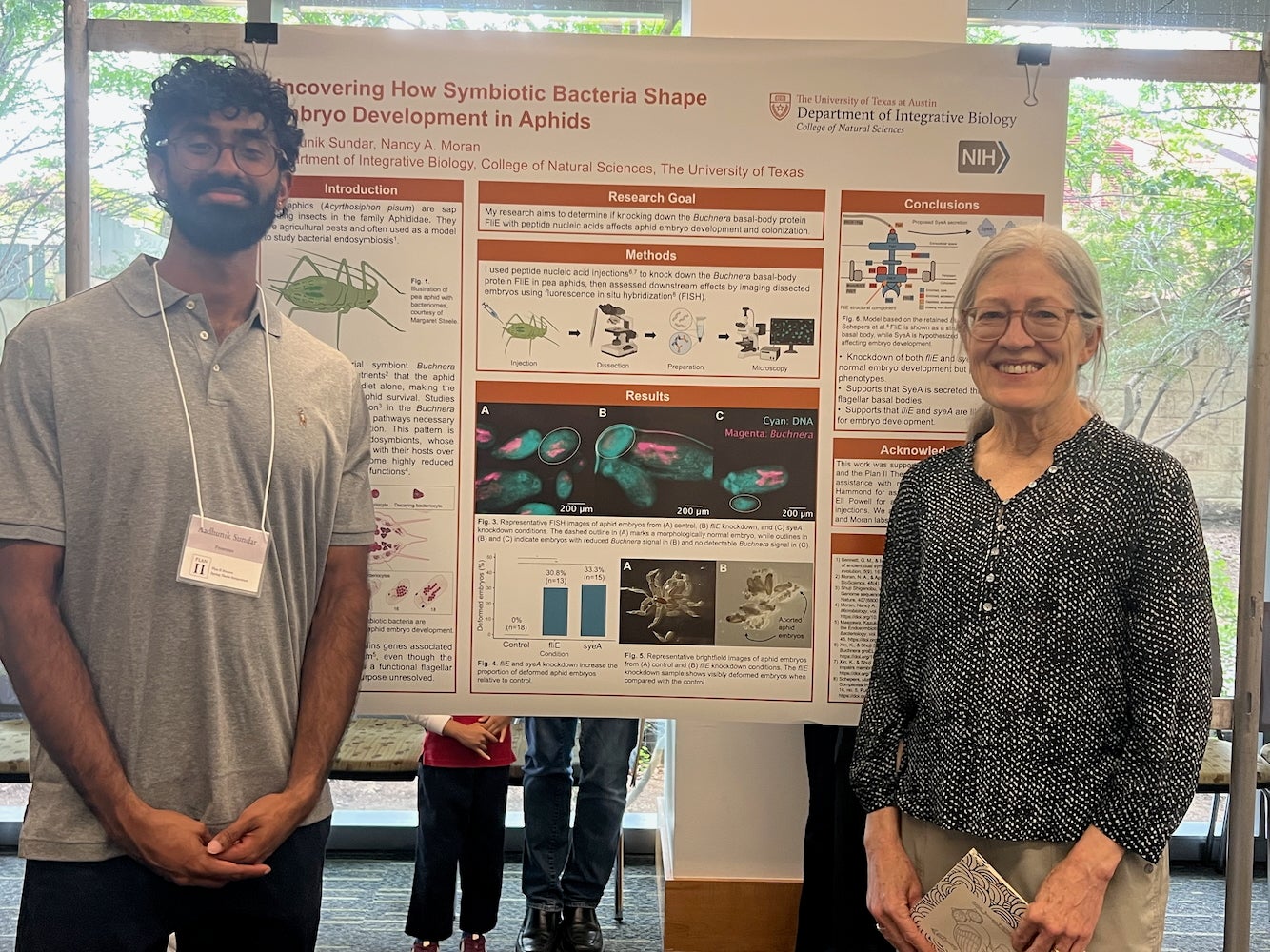 A young man and his professor pose in front of a research poster titled "Discovering how symbiotic bacteria shape embryo development in aphids."