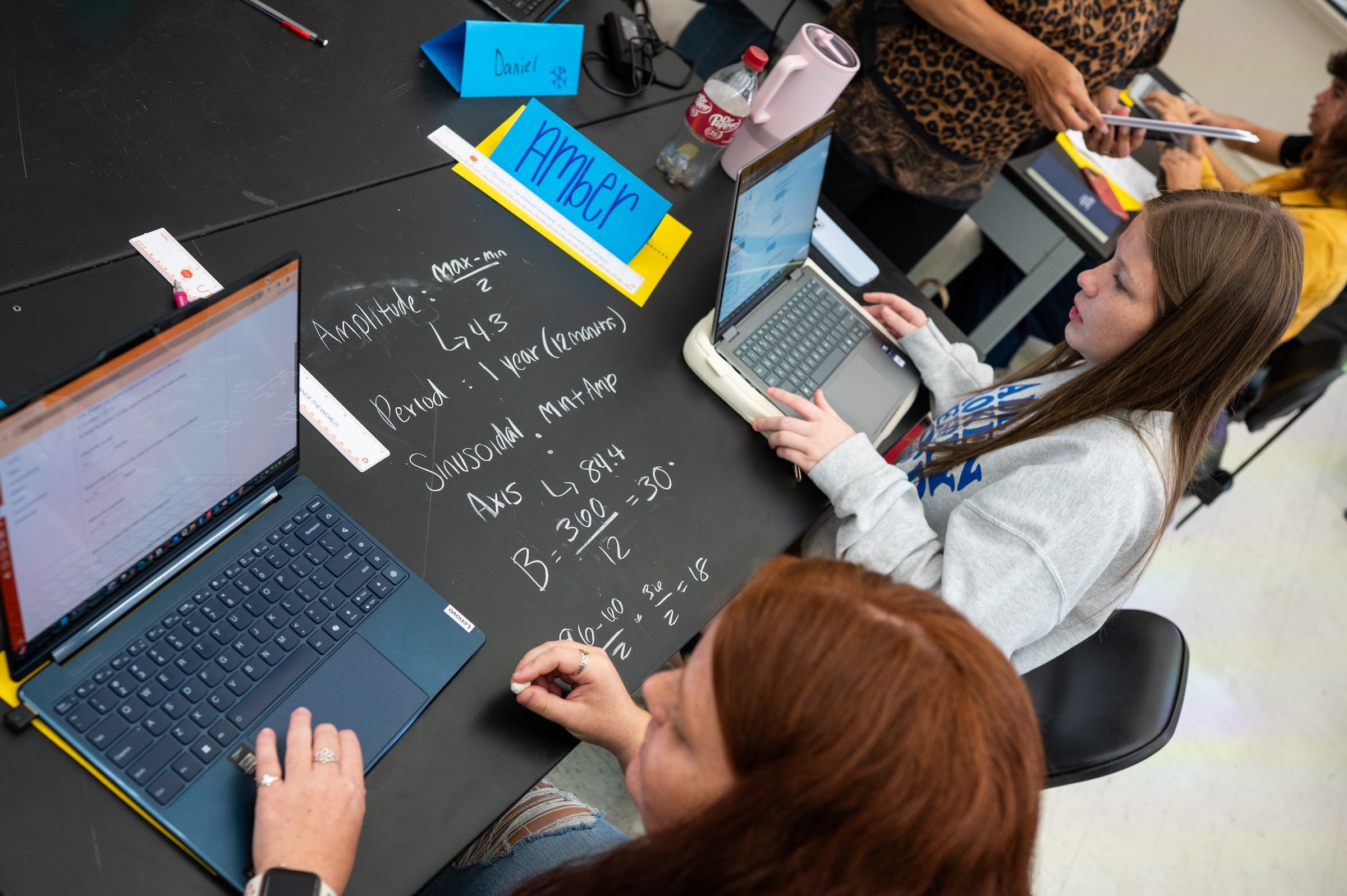 Two students at laptops flank a black tabletop with equations written on it.