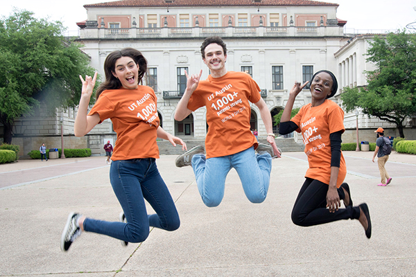 3 Students wearing t-shirts celebrating 1,000+ patents jump for joy in front of the UT Austin tower