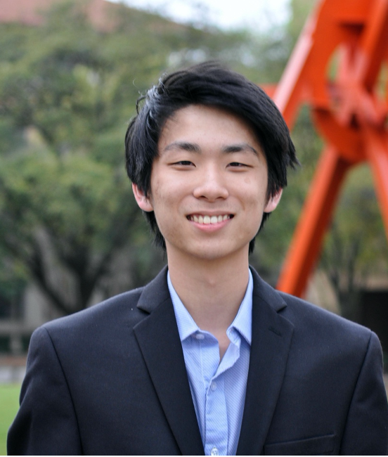 A student stands smiling in front of a Landmarks art sculpture outdoors on the UT Austin campus while wearing a collared shirt and jacket