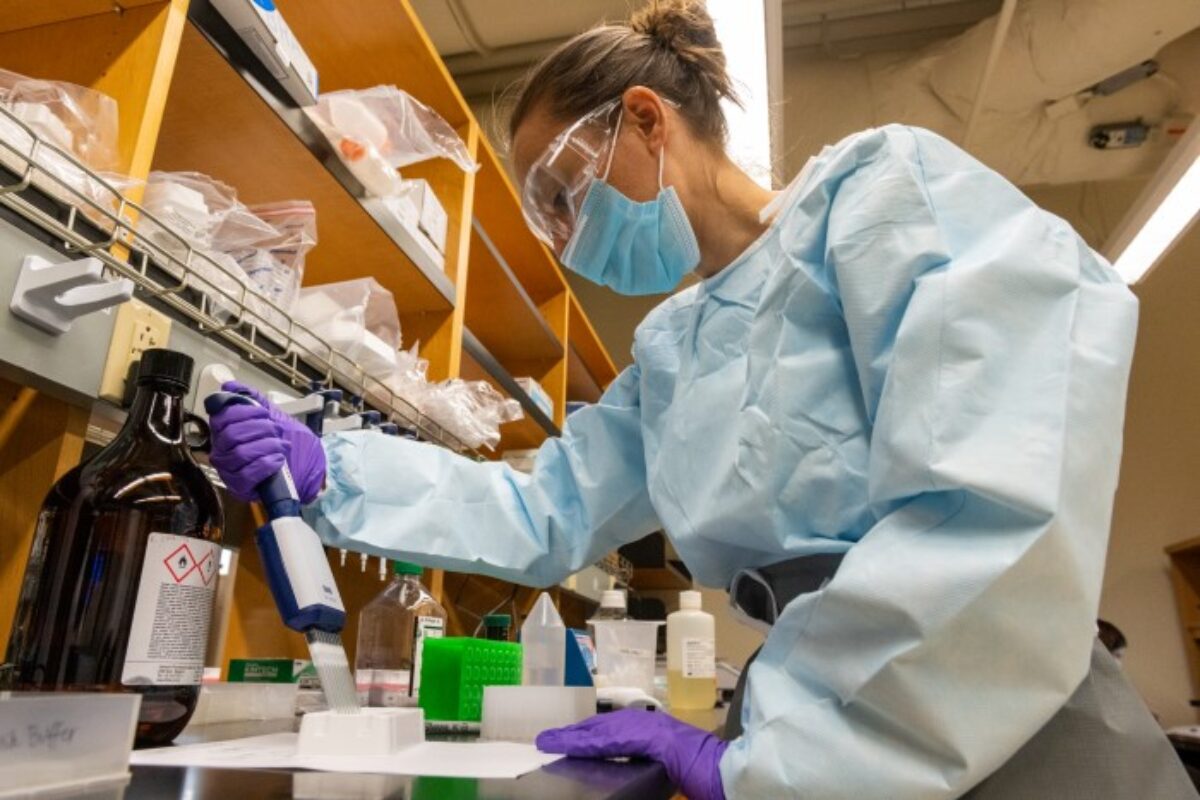 Photo shows a woman in lab protective equipment and mask and goggles handling pipette at a lab bench