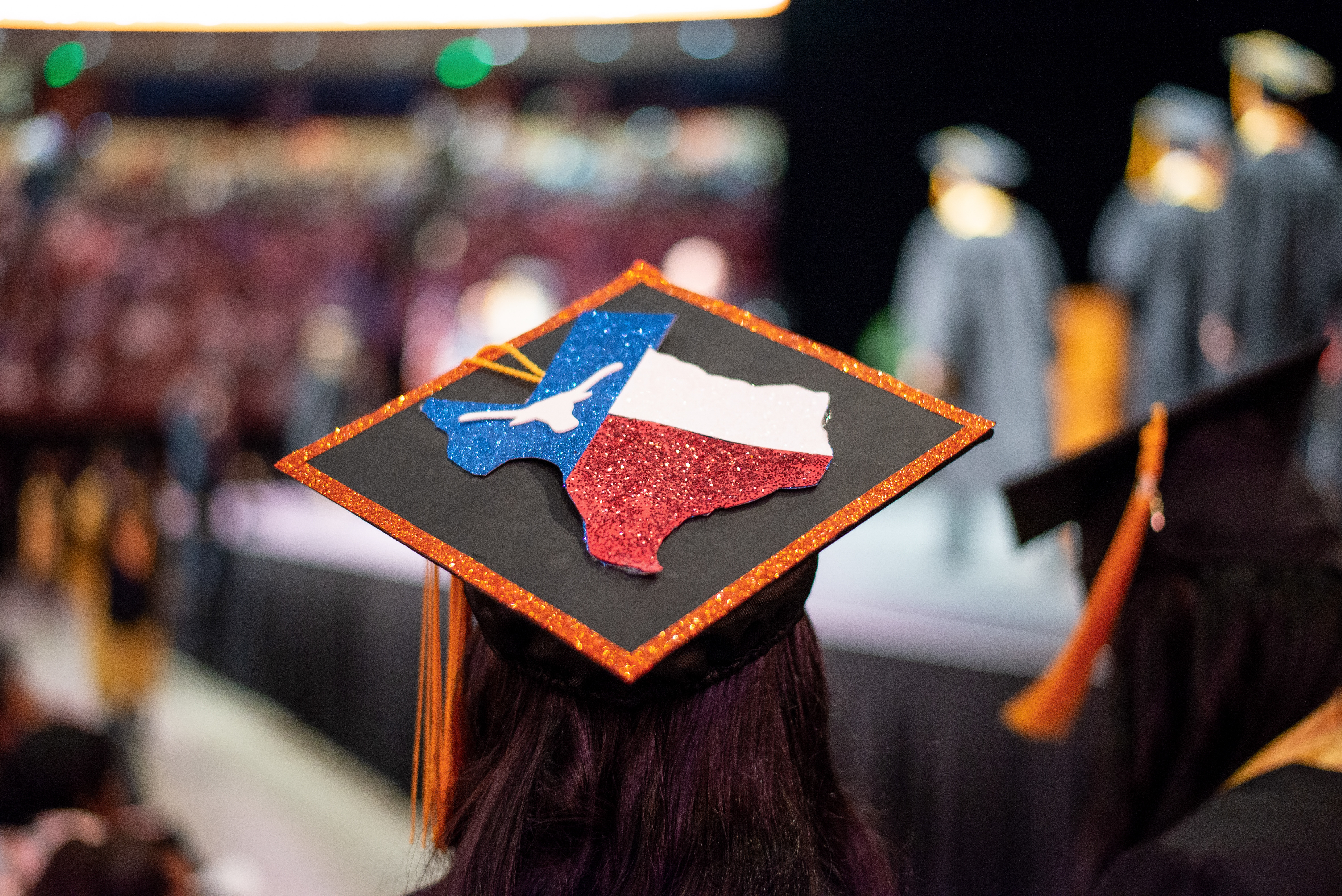 A decorated graduation cap seen from behind is lined in burnt orange with the shape of Texas and Texas flag with a UT longhorn where the star of Texas would be. The graduate is looking on as others cross the graduation stage.