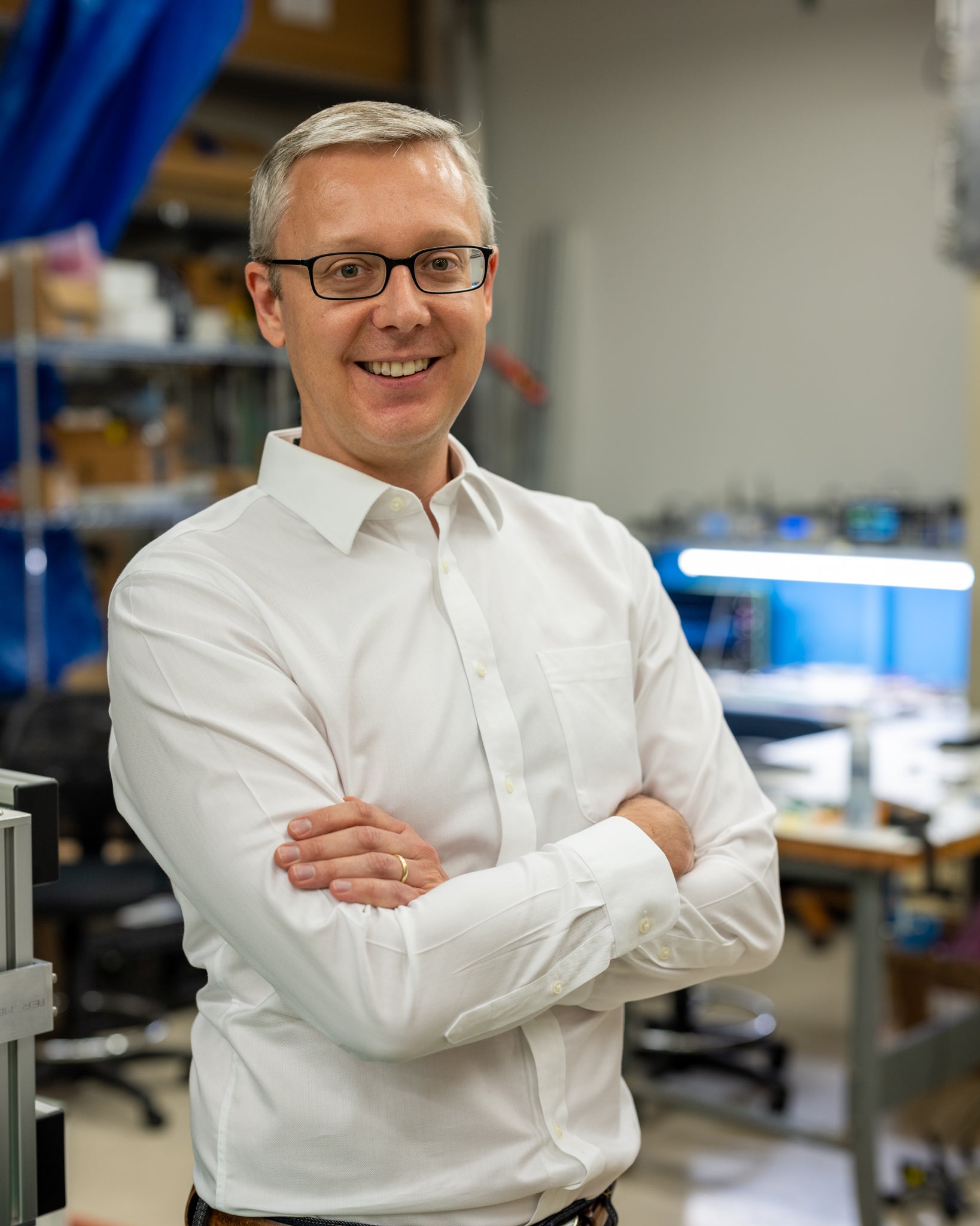A man wearing glasses and a buttoned-up shirt smiles, arms folded, while standing in a lab with equipment.