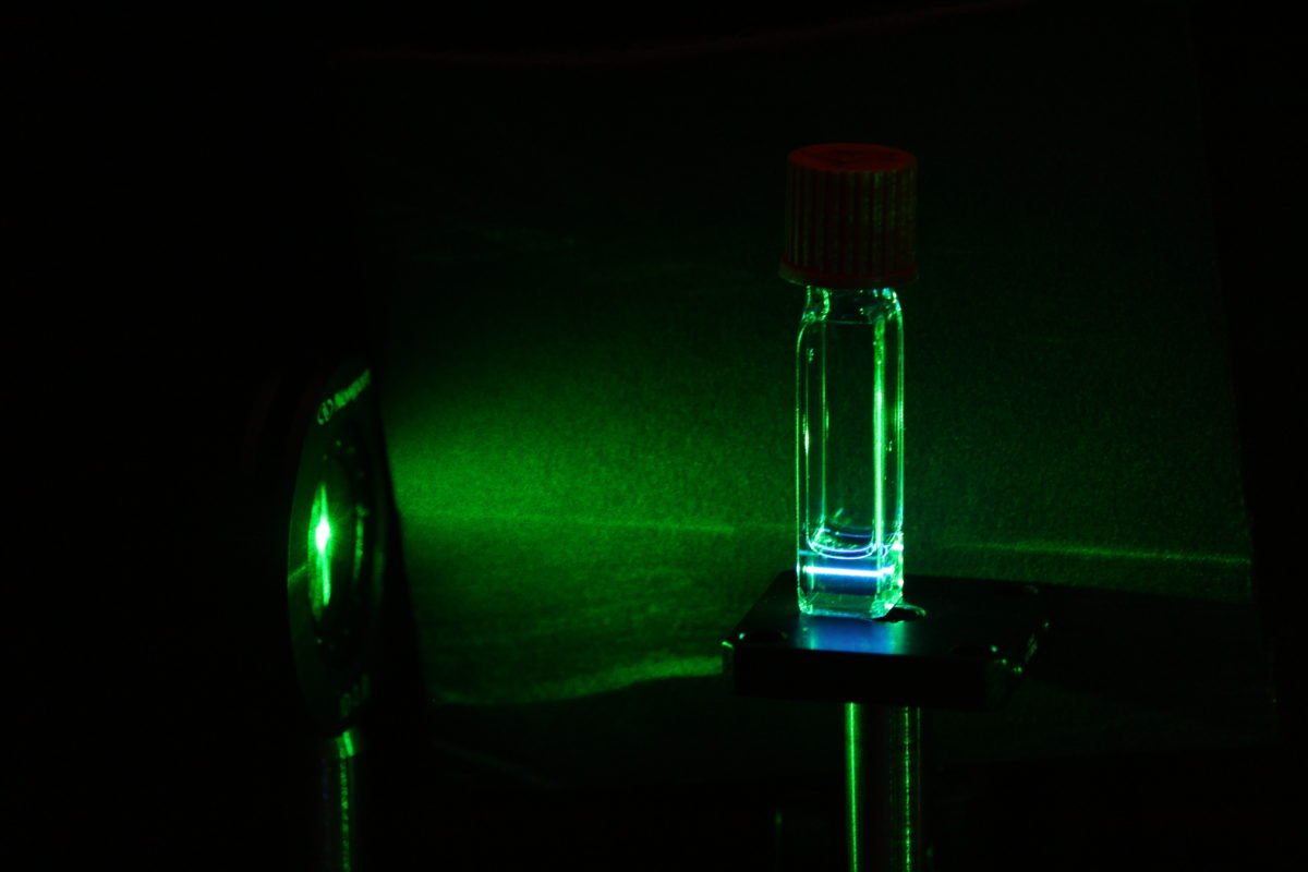 Image shows a green laser passing through a glass vial of a chemical solution
