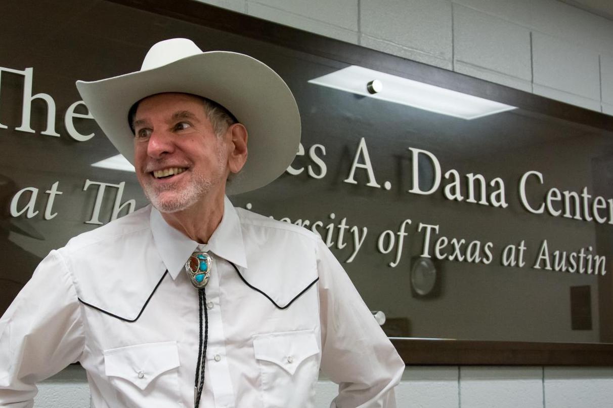 Uri Treisman wears a cowboy hat, bolo tie and Westhern shirt, smiling in front of a sign that reads The Charles A. Dana Center at The University of Texas at Austin