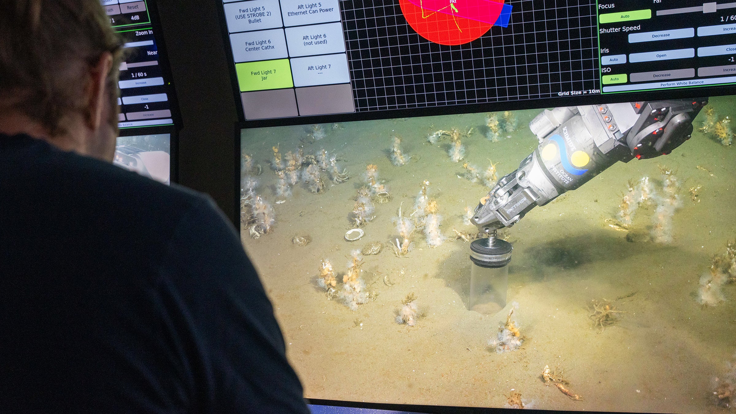 A large video monitor shows a robotic arm reaching down to the seafloor with a clear plastic tube to collect a sample of sediment