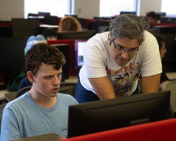 An instructor helps a student at a computer in a room where many participants are stationed at monitors.
