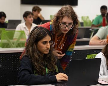 A student provides coaching to another student working at a laptop in a room of rowed participants each at their own computing station.