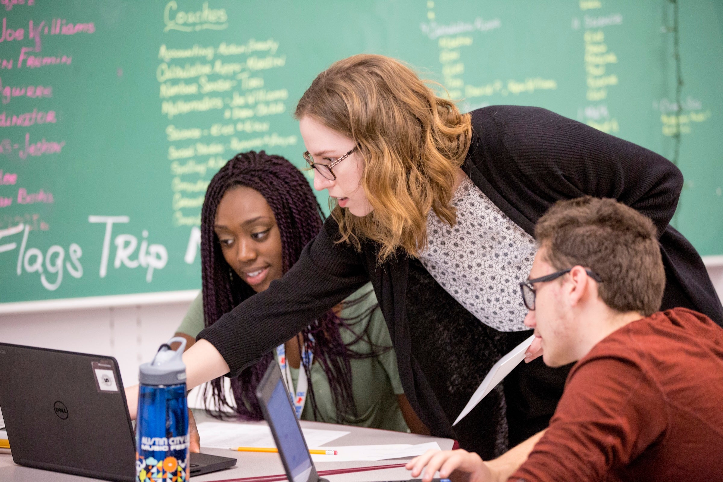 A teacher leans over a table with teen students, as the chalkboard behind her has lists of words and phrases.