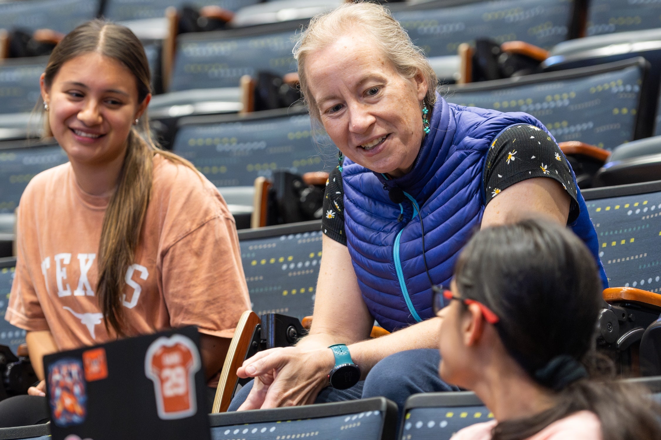 Two students in rows chat with an instructor after class, who leans in with a smile.