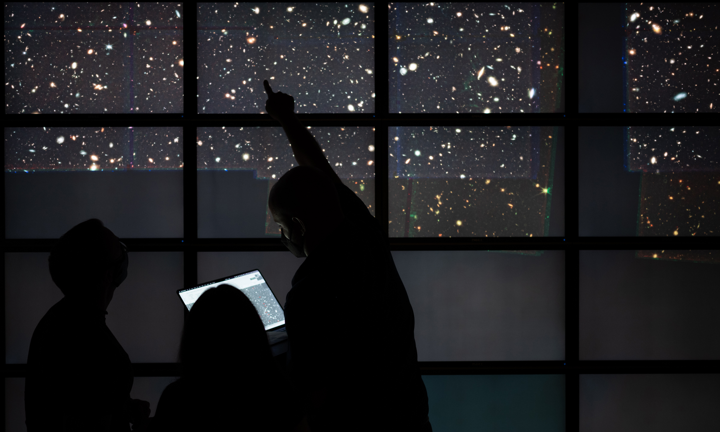 Silhouettes of three people in front of a large display of stars and galaxies, one person pointing at a specific spot