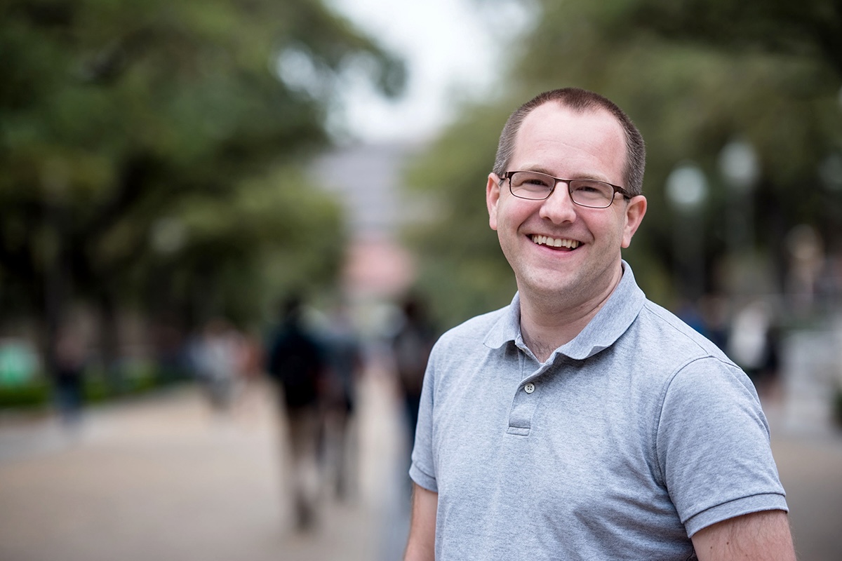 A man in glasses and a polo shirt smiles while outdoors on a campus with people passing by in the distance