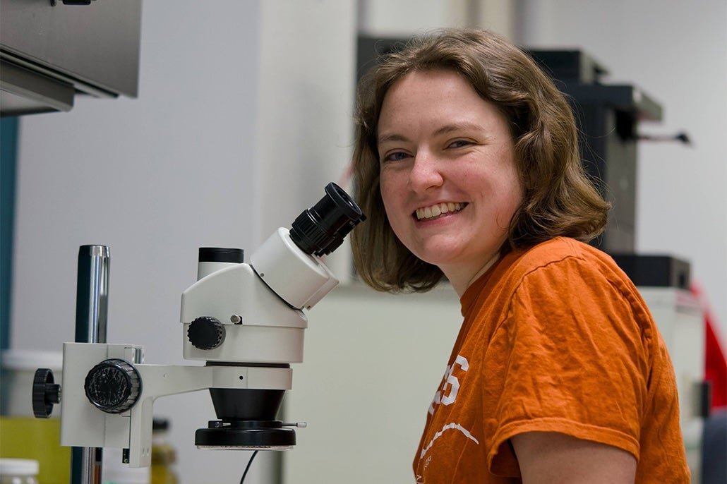 A young woman in burnt orange smiles while sitting in front of a microscope in a lab and collections space.