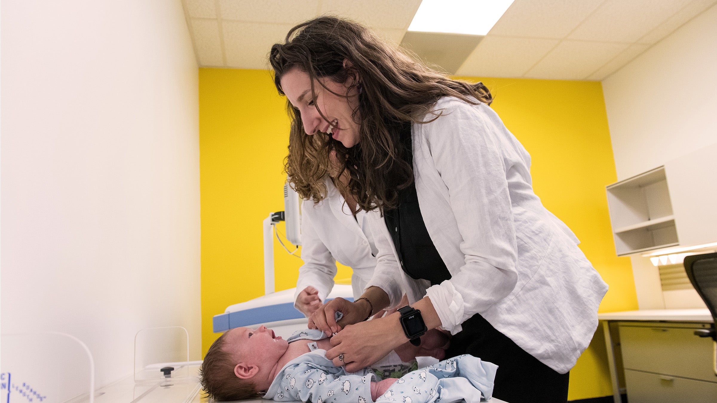 Beth Widen, a dark haired woman in a white coat, leans over an infant lying on a table. She smiles down at the baby. 