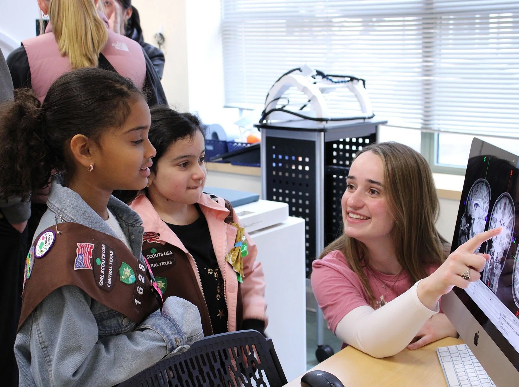 Two elementary-aged girl scouts marvel at images of a brain on screen, as an undergraduate student points to a label on the image.